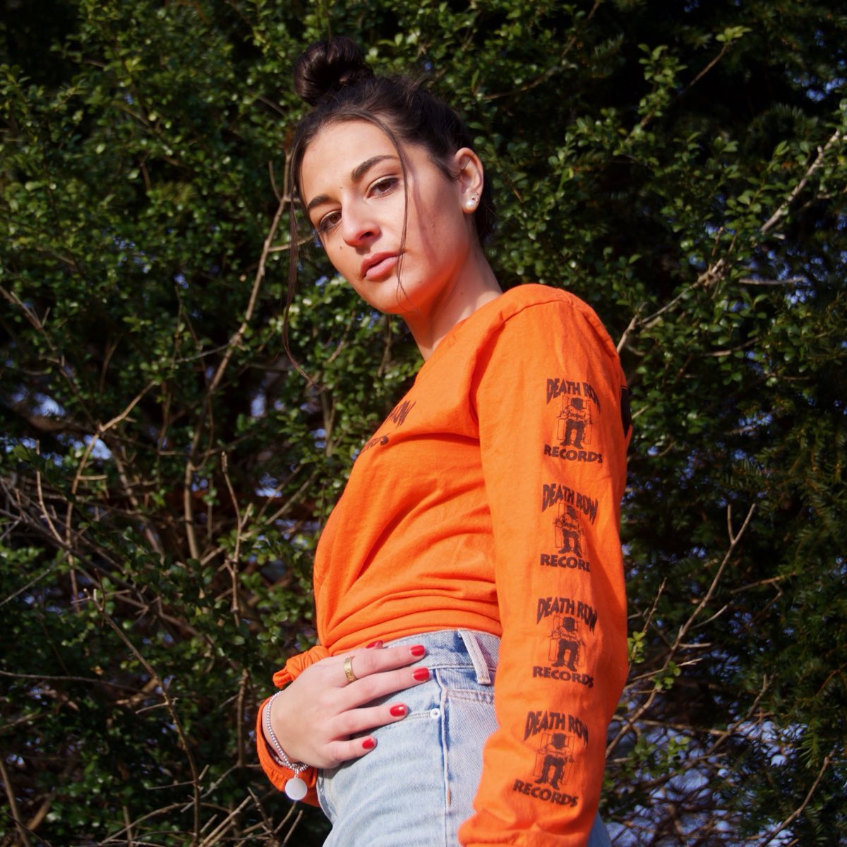 Portrait of a woman in an orange long-sleeve top, backlit against summer trees.