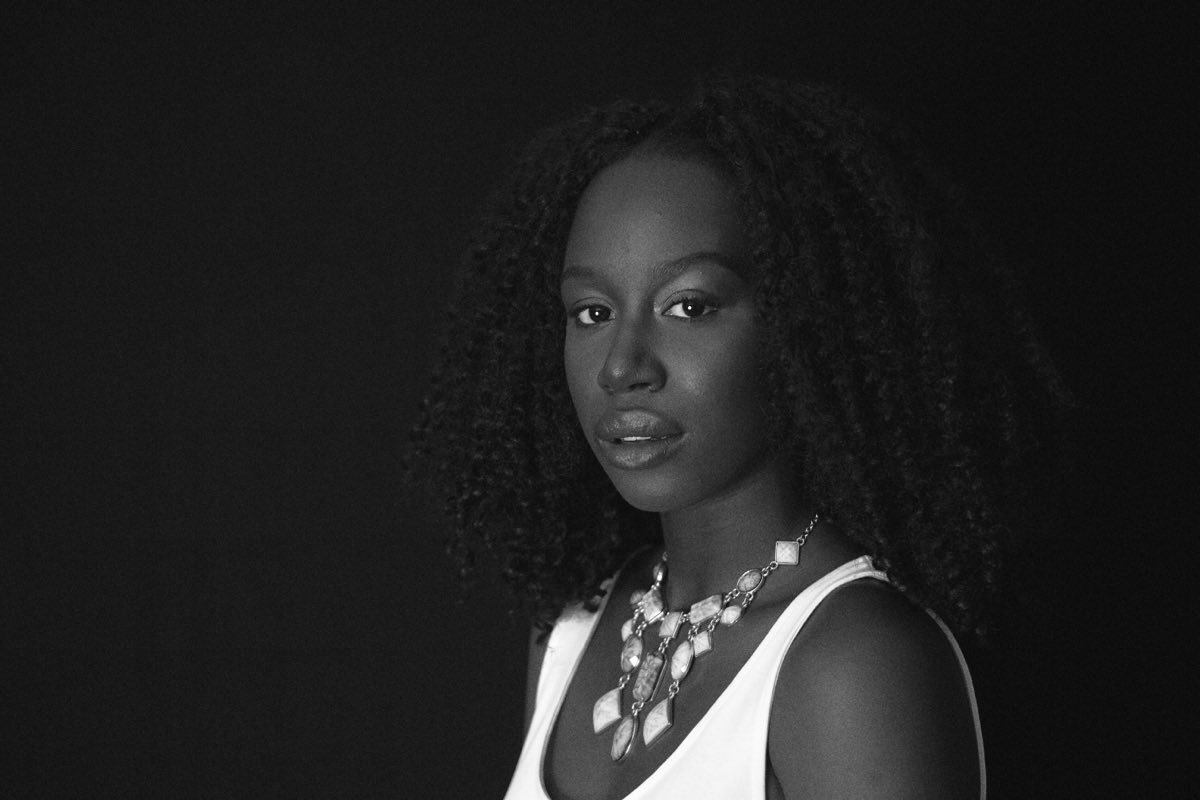 Black and white studio portrait of a woman against a dark ground, looking straight to camera.