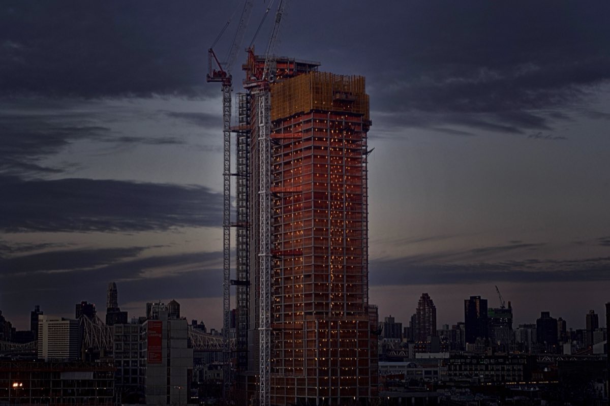 A tower under construction lit red against the New York skyline at dusk.
