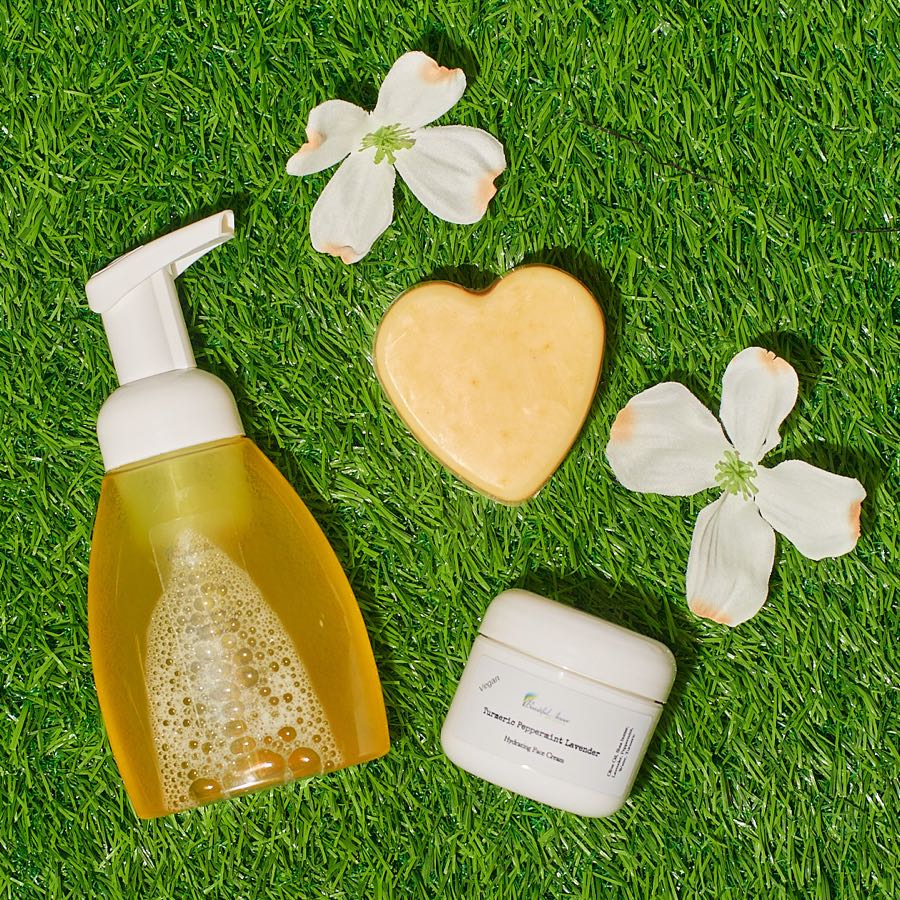 Beautiful Amore foaming body wash, a heart-shaped soap and a jar of cream arranged on grass with dogwood blossoms.