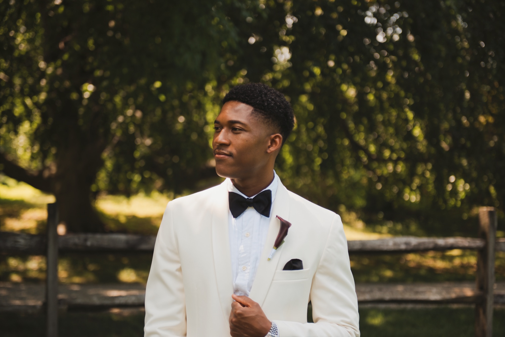 The groom in a white dinner jacket and black bow tie, looking off to the side under a weeping tree.