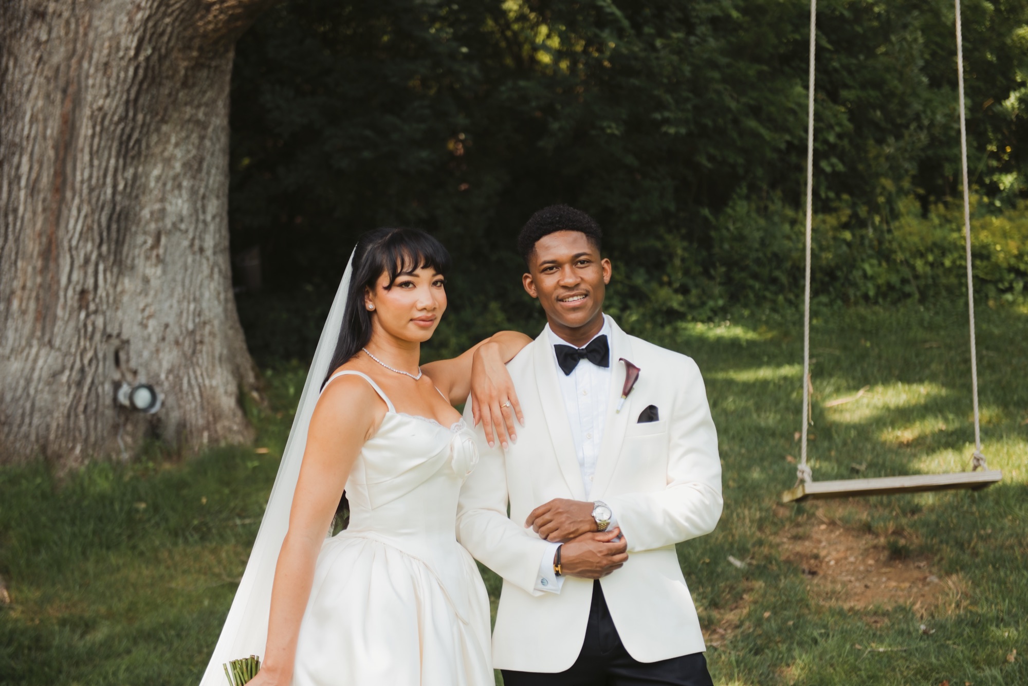 The bride resting her arm on the groom's shoulder beside a rope swing, both looking at the camera.