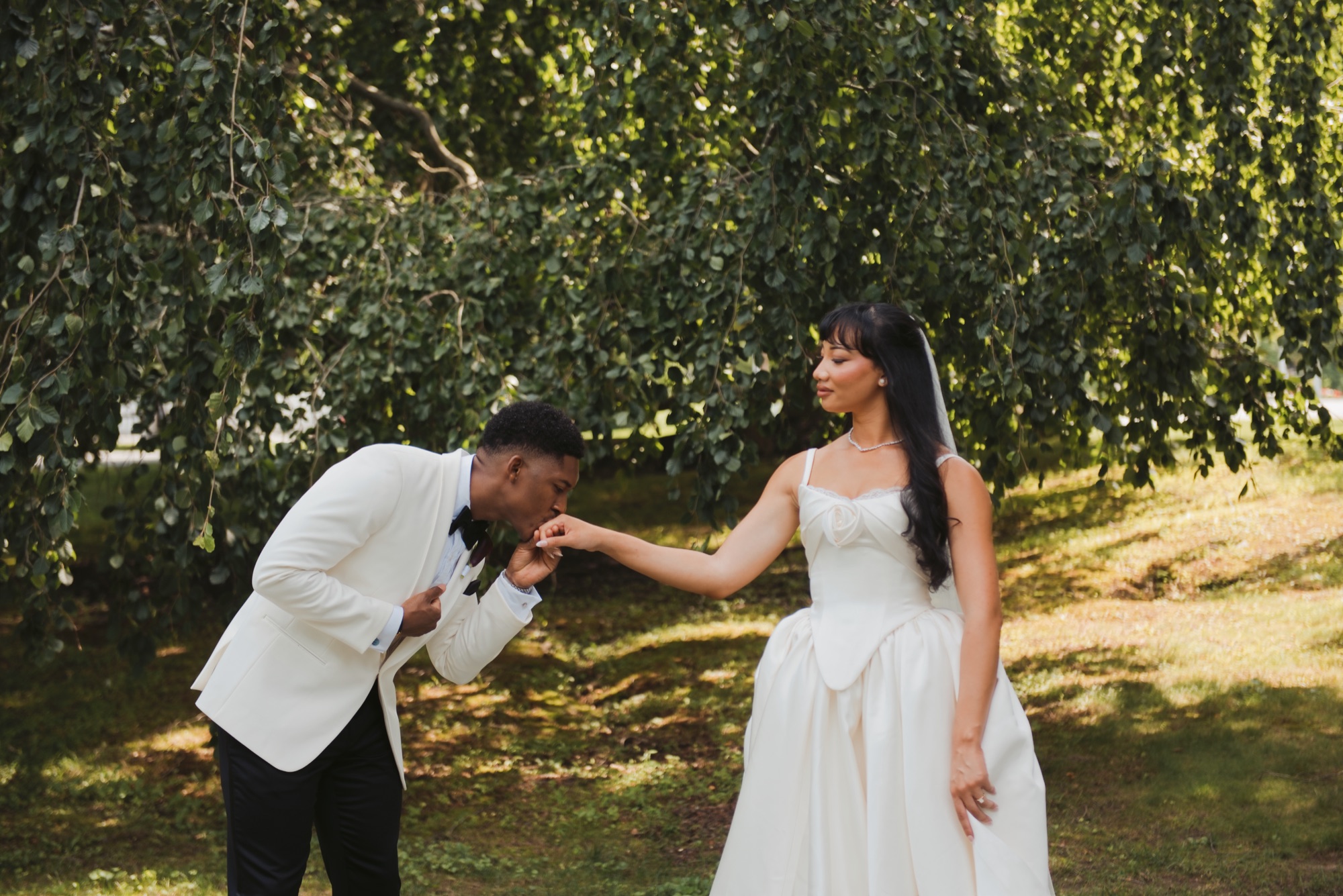 The groom bending to kiss the bride's hand in dappled light.